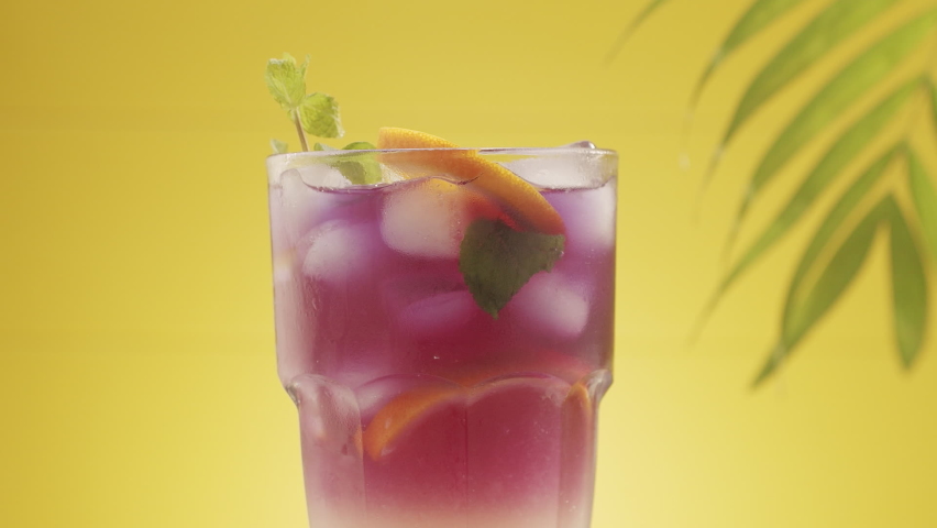 Glass of refreshing summer lemonade cocktail with ice and lime and mint rotating, Summer refreshing cocktail, studio shot on yellow background with tropical leaves with sea sand. Citrus Lime Cocktail.