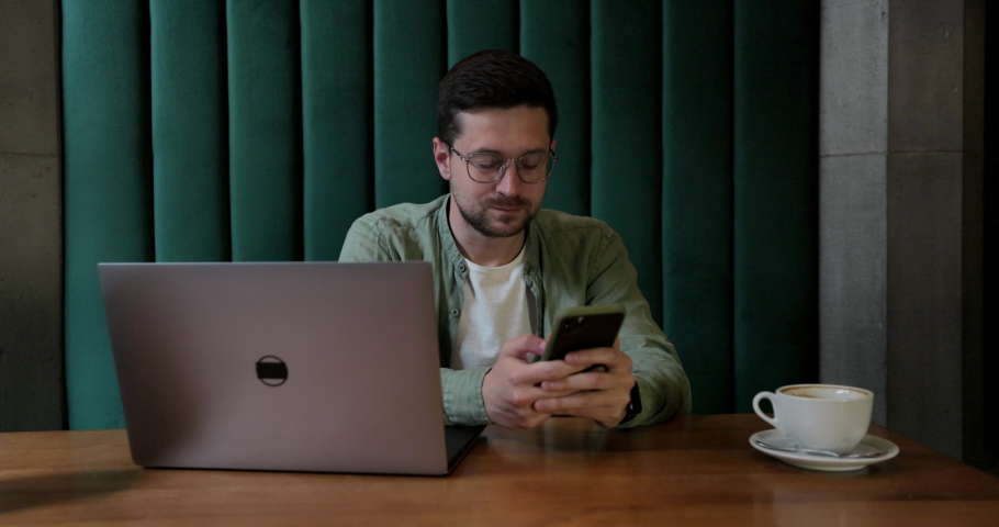 Young man in glases typing on smartphone in coffee shop, laptop on the table. Man checking information on cellphone chatting on phone in cafe.