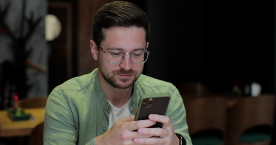Young man in glases and casual wear sitting with smartphone in the hands, browsing something, working at the cafe. Man is using smartphone for messaging, texting.