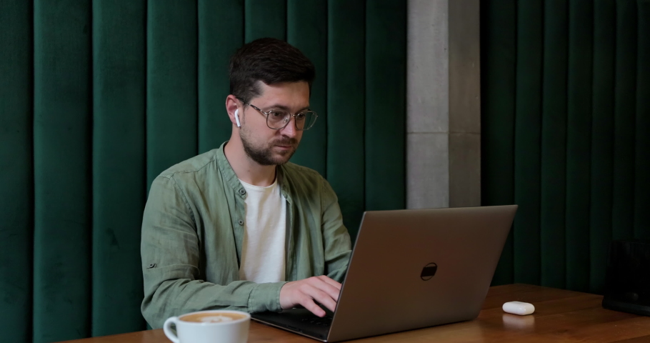 Man in glases typing on keyboard of laptop while sitting at cafe table. Young guy using portable computer for remote work.