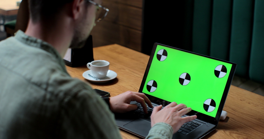 Young man in glases working on laptop with green screen in coffee shop.