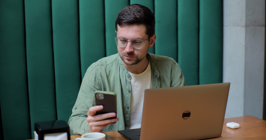 Young man in glases working on laptop and checking smartphone for messages in small cafe.