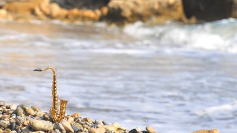 Beautiful golden alt tenor saxophone stands against backdrop of blurry sea amid raging waves. Music screensaver romance. Copy space for your text. Mini model real saxophone. Slow motion video.
