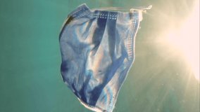 VERTICAL VIDEO: Slow motion, used medical face mask slowly swim underwater on sunrise time in sunbeams. Backlight (Contre-jour). Discarded face masks polluting seas since COVID-19
