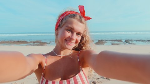 Young positive Caucasian woman teenager films himself on phone sending air kiss sends greetings to friends or boyfriend enjoys seaside vacation stands on sand of sunny tropical beach. Camera view