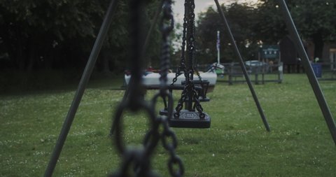 HANDHELD SHOT of a defocused swing chain in the foreground and focused swings in the background, swinging slowly at dusk. SLOW MOTION, BOKEH.