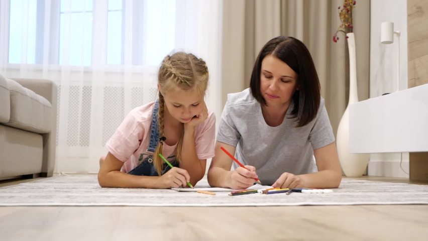 Smiling young mom and cute preteen daughter lying on warm floor at home drawing in album together, happy parent or nanny have fun spending time painting with colorful pencils, slow motion