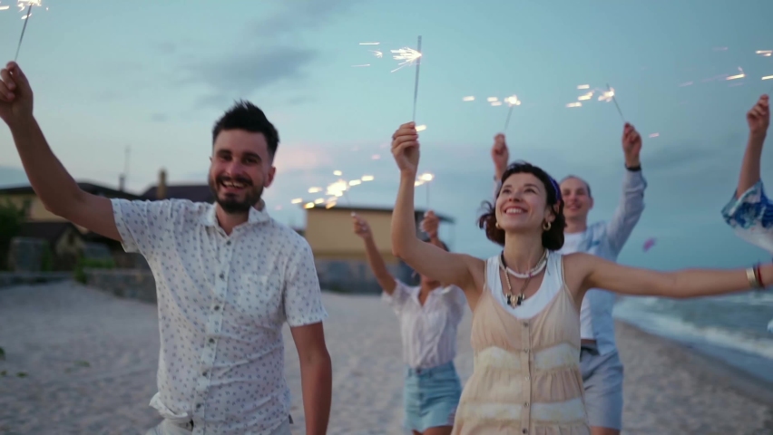 Multiethnic friends walking, dancing, having fun on night party at seaside with sparklers lights in hands on Fourth of July. Young people partying on the beach with fireworks on Christmas or New Year. | Shutterstock HD Video #1091410399