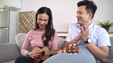Asian couples are happily playing ukulele together. Men are singing and clapping while women are playfully playing the ukulele on the sofa in the living room in couple and lifestyle concept