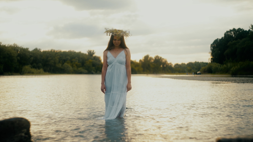 Magical beautiful girl of Ukrainian nationality with black hair with closed eyes stands at sunset in a white dress in the water. Opens eyes in slow motion. Artistic effect, soft focus. lights shine.