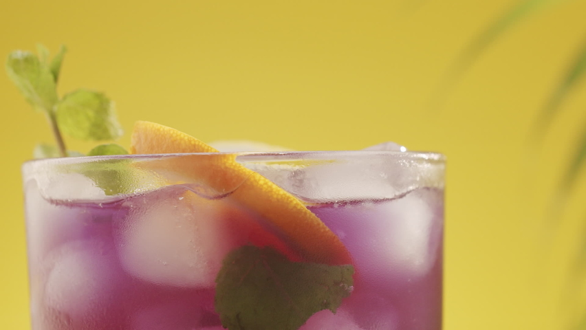 Summer refreshing cocktail, studio shot on yellow background with tropical leaves with sea sand. Citrus Lime Cocktail. Glass of refreshing summer lemonade cocktail with ice and lime and mint rotating.