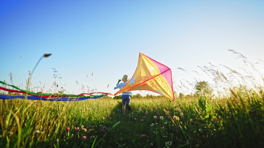 Fast little boy runs across the field with a kite in his hands fluttering in wind over his head. Front view on child walks in nature in summer and has fun play. Family trip out of town to fresh air. Royalty-Free Stock Footage #1091420629
