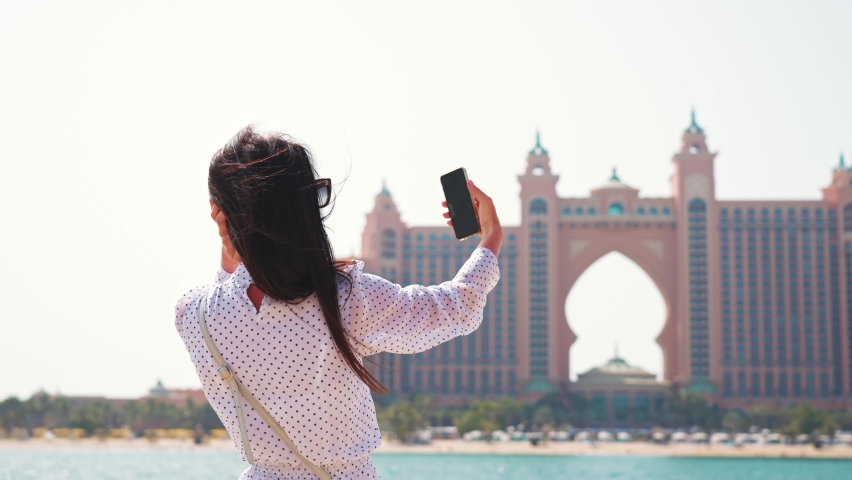Girl holds phone in her hand and takes selfies against the sea and luxury 5 star hotel on artificial island in Dubai (UAE). Young woman on vacation and enjoy weekend at resort.