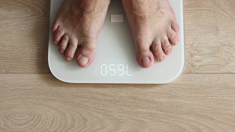 Male bare feet stepping on white digital floor scales - man weighing himself at home: close up top view. Measuring weight, technology, control, wellness and diet concept.