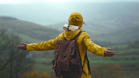 Beautiful happy smiling woman in yellow raincoat, retro backpack and knitted beanie stands outdoors against fields, raises arms, inhale and breathe in fresh rain air. Travel adventure tourism concept. - Powered by Shutterstock - Get 15% off with code: PIKWIZARD15