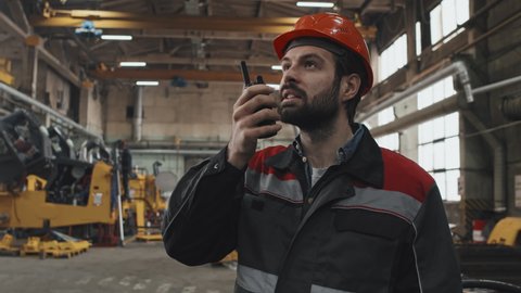 Medium close-up of Caucasian bearded male plant manager talking into walkie talkie at factory in afternoon