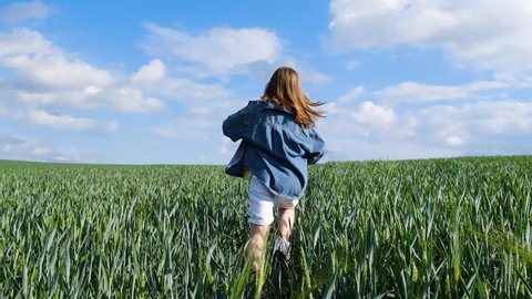 A teenage girl in denim clothes runs across a wide green field. A child is playing in a wheat field