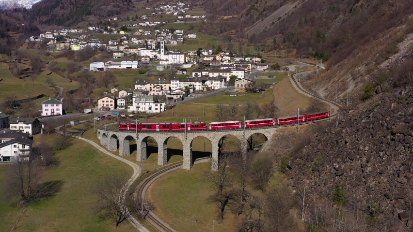 Train on Brusio Spiral Viaduct in Switzerland on Sunny Day. Bernina Railway. Swiss Alps. Aerial View. Drone Flies Backwards and Upwards