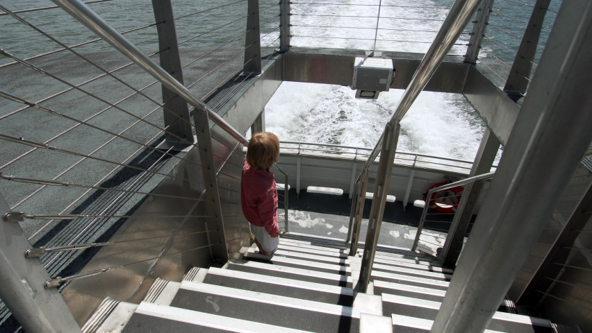 A 5 years old caucasian boy in black mask is looking at the foamy waves on water surface behind of fast moving NYC ferry. Travelling by NYC ferry. Summer day. USA