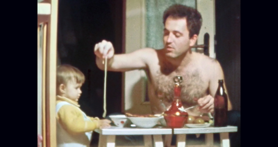 Funny dad with child at table with food in home room. Happy, smiling father invites little child eat food. Parent having fun. Vintage color film. Family humor. Life archive. Retro 1980s Kyiv, Ukraine