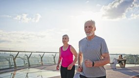 Positive senior man and slim mature woman run on city panoramic observation deck at sunrise. Pensioner checks time looking at smartwatch on wrist slow motion - Powered by Shutterstock - Get 15% off with code: PIKWIZARD15
