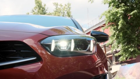 close view of front headlight of modern orange colored car. headlight glows with diode lights in new 2022 model car. unrecognizable car is parked in alley.