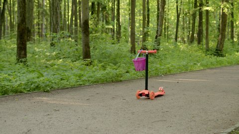 small child girl with ponytail runs up to orange scooter in park, takes it and leaves frame. cheerful little girl in striped t-shirt and black pants driving scooter rolls through forest along path.