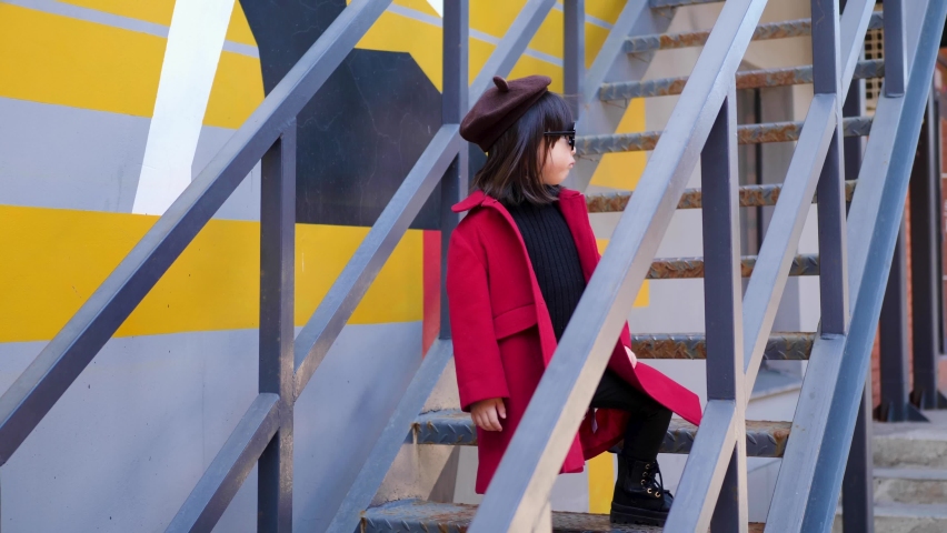 Korean girl in a red coat and cap and round glasses sit on the iron staircase the street in autumn.