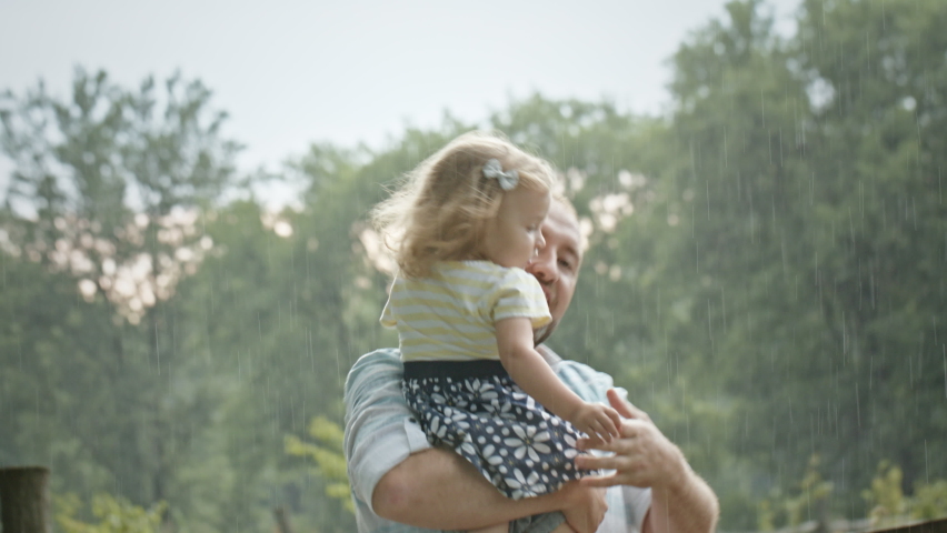 Father and daughter dancing under rain. Happy adult dad holding daughter in arms and kissing hand of kid while spinning around near wooden fence and enjoying rain on summer day in countryside Royalty-Free Stock Footage #1091460637