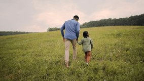 Rear of African American man holding hands with cute small son walking on meadow in grass and talking. Dad and kid spending leisure time together, green field, nature, rural area, family concept - Powered by Shutterstock - Get 15% off with code: PIKWIZARD15