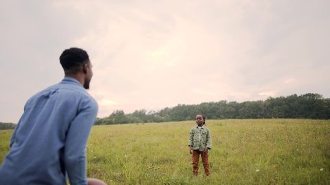 Rear of African American father playing with little son kid with ball standing in field. Cute child boy plays american football throwing and catching ball in nature. Happy childhood concept