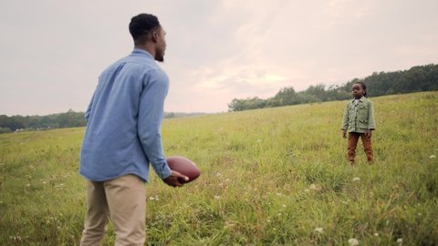 African American caring father playing with little son kid throw and catch with ball standing in field outdoors. Dad teaching child to play american football in meadow. Happy childhood. Family concept