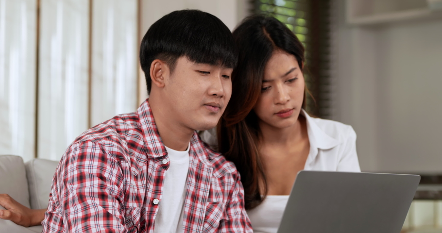 Slow motion shot, Happy Young couple sitting on sofa in living room at home use laptop computer working, talking and smile together with happiness, happy family concept