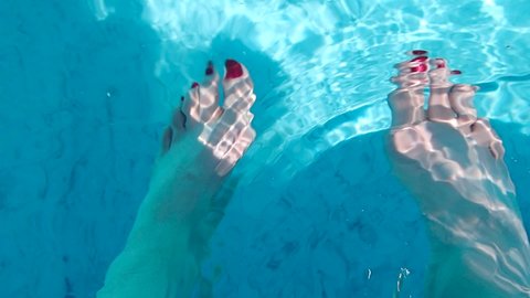Clean blue swimming pool water sparkles in the sun as it ripples and flows over a woman's feet with red nail polish that move up and down in the water in unison. High energy fun vacation footage