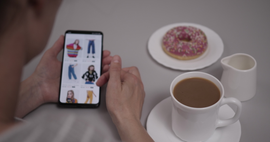 Woman Looks at Goods in Online Store Clothing. Buy Fashion Clothes Directly on Smartphone. Woman at Home Sitting at Table With Coffee and Donut Using Smartphone Buys in Internet Shop. Royalty-Free Stock Footage #1091472599
