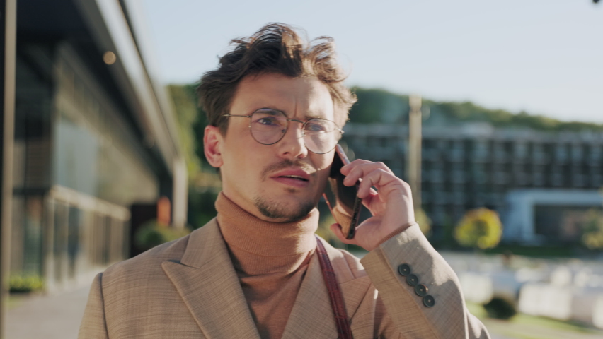 Close up face serious young man in glasses speak on phone look around walking at street. Modern architecture. Sunlight. Slow motion