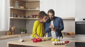 Happy caucasian couple smiling cheerfully while looking on screen of modern smartphone while standing together on kitchen. Man and woman watching funny videos and cooking fresh lunch.