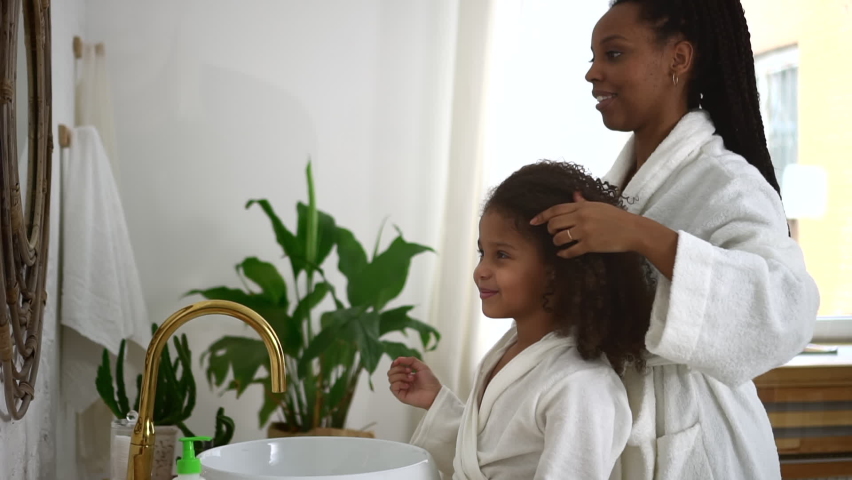 Young mother, daughter are doing daily hygiene and standing in home bathroom spbd. Close view of american african woman, cute girl cleanse body and look in mirror with smiles, have fun and stand in