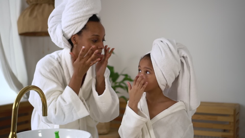 Mom, daughter are doing beauty treatment and smiling while standing in home bathroom spbd. Close view of american african woman, girl apply cream or mask on face skin and look with smiles. Royalty-Free Stock Footage #1091484357