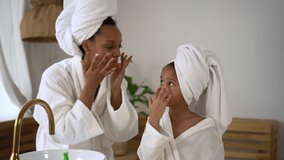 Mom, daughter are doing beauty treatment and smiling while standing in home bathroom spbd. Close view of american african woman, girl apply cream or mask on face skin and look with smiles. - Powered by Shutterstock - Get 15% off with code: PIKWIZARD15
