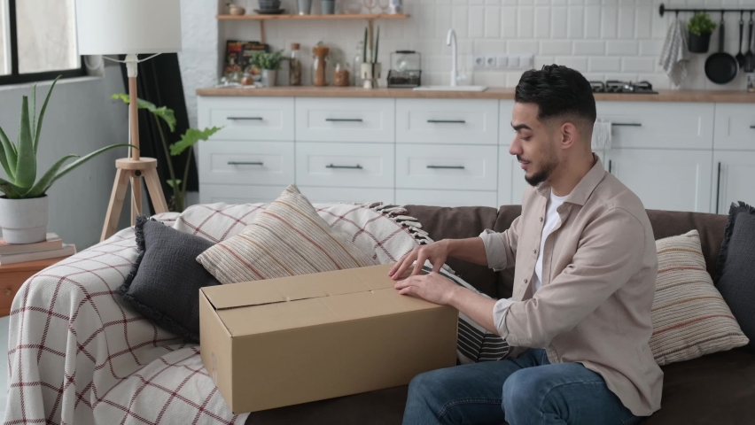 Happy attractive indian or arabian guy opening big carton box, unpacking internet store order at home, contented with fast delivery service and the goods which received, smiling and feel satisfied
