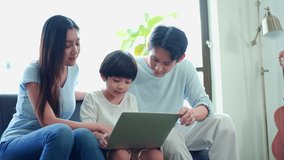 Happy family, Asian young son with mom and dad sitting on sofa together. while using laptop computer browsing online internet watching cartoon, social media enjoy during holiday at home.