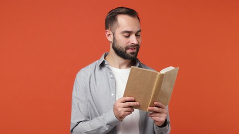 Smiling fancy amazing swanky young brunet man 20s years old wear blue shirt reading book isolated on plain orange background studio portrait. Education high school collage university free time concept