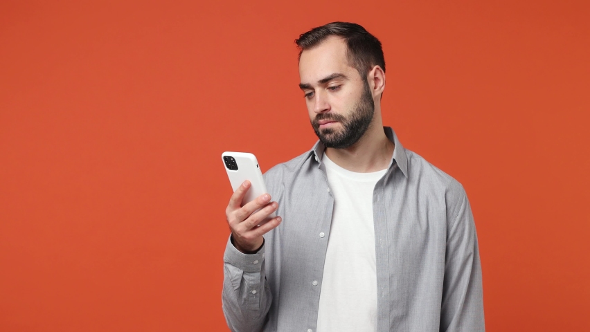 Excited joyful overjoyed jubilant young brunet man 20s years old wears blue shirt using mobile cell phone hold fan of cash money in dollar banknotes isolated on plain orange background studio portrait