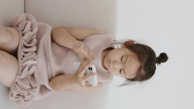 Portrait of a cute attractive charming sweet calm peaceful focused Asian girl using the 5g online internet application in a light white interior of the living room of the house indoors, vertical video