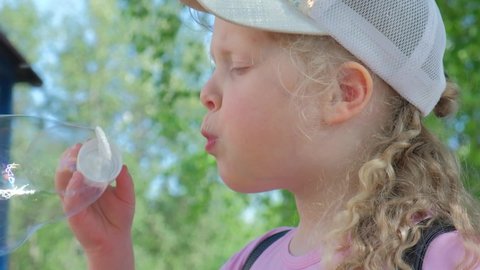 cute girl 6 years old inflates bubbles in nature, the child has fun with soap bubbles, blond girl preschooler in a cap slow motion