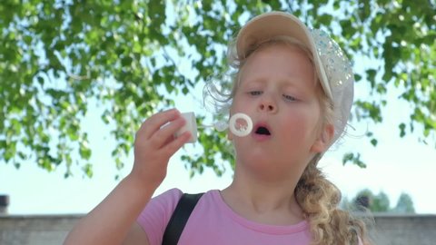 cute girl 6 years old inflates bubbles in nature, the child has fun with soap bubbles, blond girl preschooler in a cap slow motion