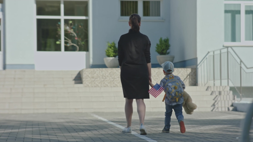 Mother in black holding preschooler boy child hand taking him back to school. backside view of kid elementary age walking with yellow backpack, beige teddy bear, going school for the first time day. | Shutterstock HD Video #1091500075