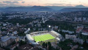 Cityscape featuring a stadium with green grass soccer field (football pitch) illuminated by floodlights at night, in the dark city of Podgorica, Montenegro, under colorful sunset sky in the evening. - Powered by Shutterstock - Get 15% off with code: PIKWIZARD15