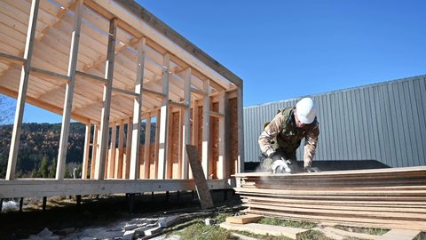 Carpenter using circular saw for cutting wooden OSB board. Man worker building wooden frame house on pile foundation. Carpentry concept.